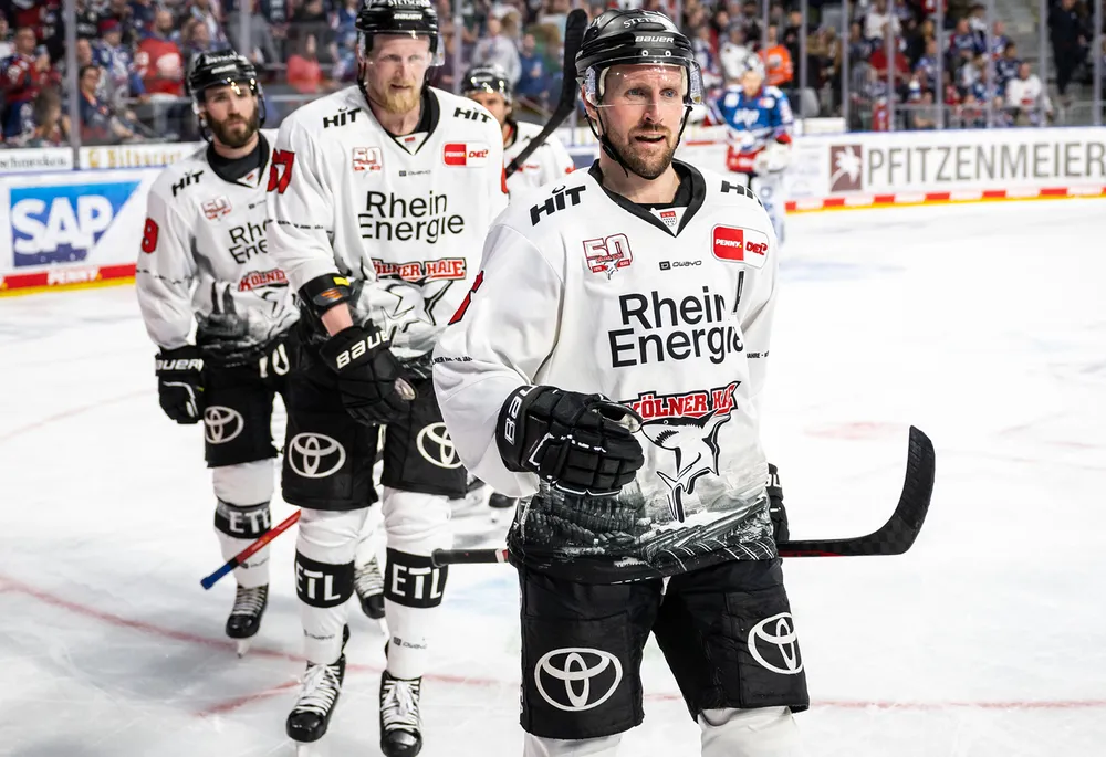 Ice hockey players in white jerseys with black pants featuring a team's logo, standing on the ice.