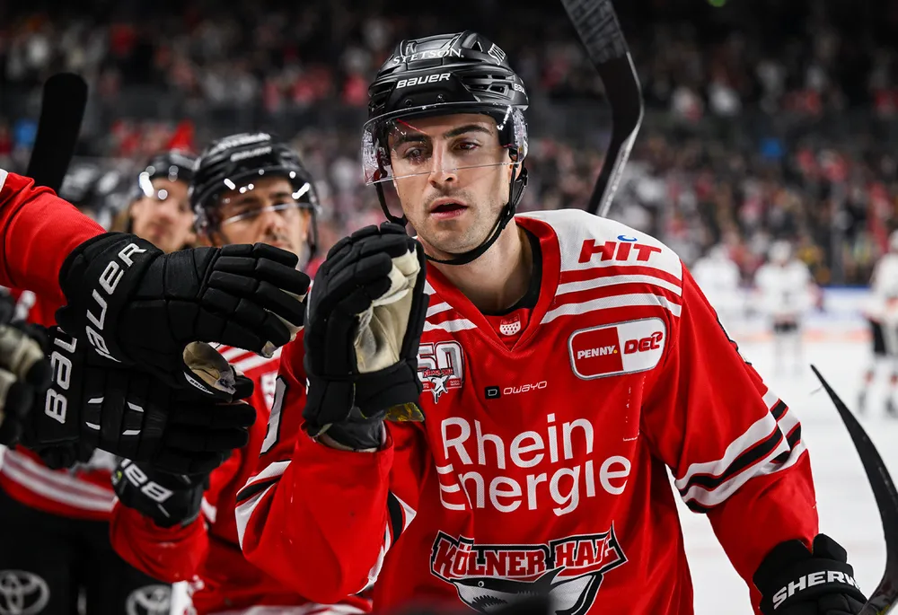 An ice hockey player in a red jersey with black gloves and a helmet, celebrating with teammates.