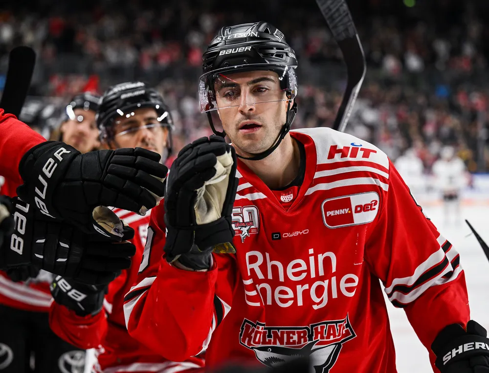 A hockey player is wearing a red jersey with white and black details while interacting with a teammate.
