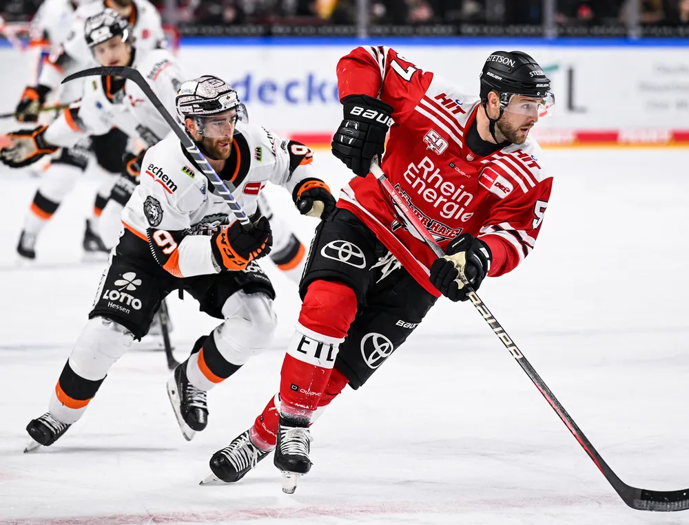 An ice hockey player in a red jersey and black pants skates on the ice while being pursued by another player.