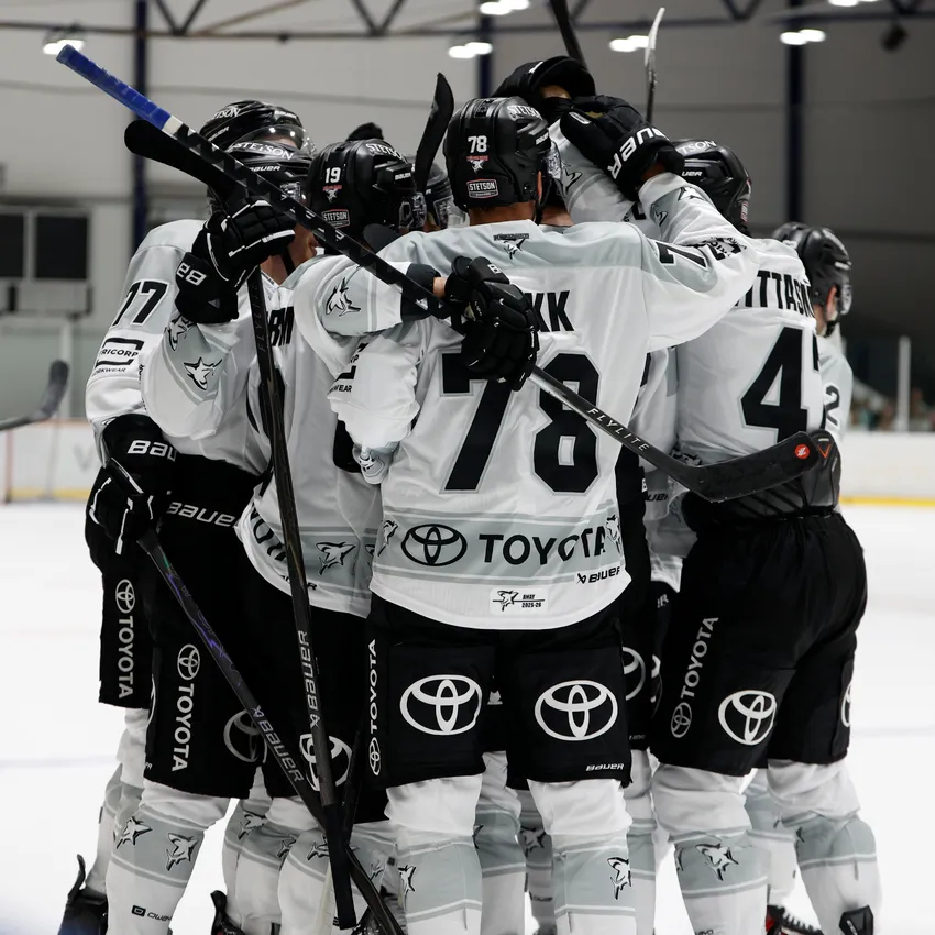 A group of ice hockey players in white jerseys embrace on the ice after scoring a goal.