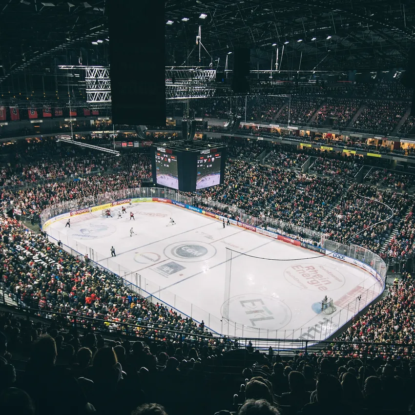 A packed ice hockey arena with cheering fans, a visible rink, and a large screen in the background.