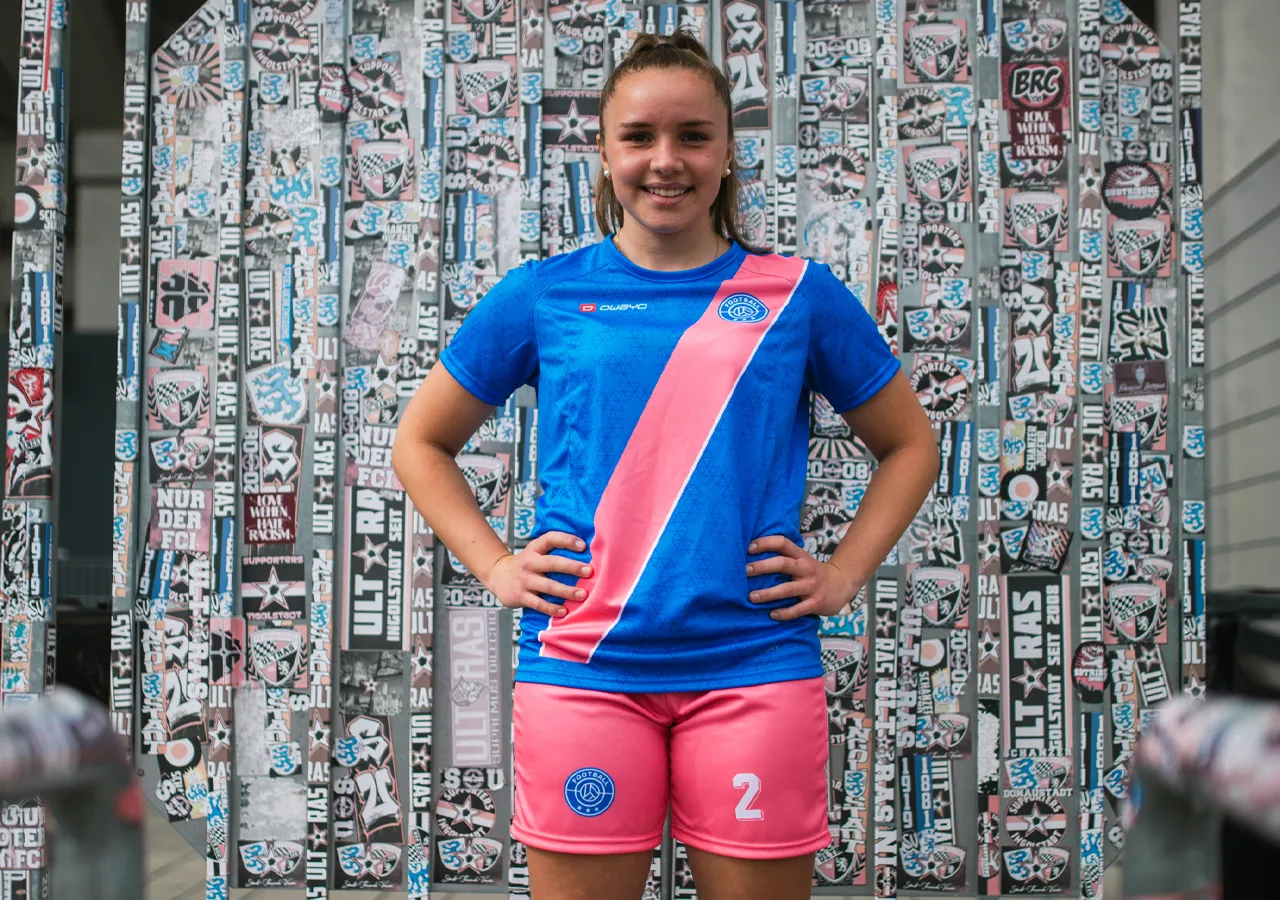 A young woman is wearing a blue soccer jersey with a pink diagonal stripe and pink shorts featuring the number 2, in front of a wall covered with graphics and stickers.