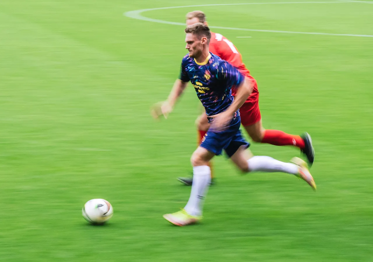 A soccer player in a colorful jersey sprints across the grass while another player closely follows behind.