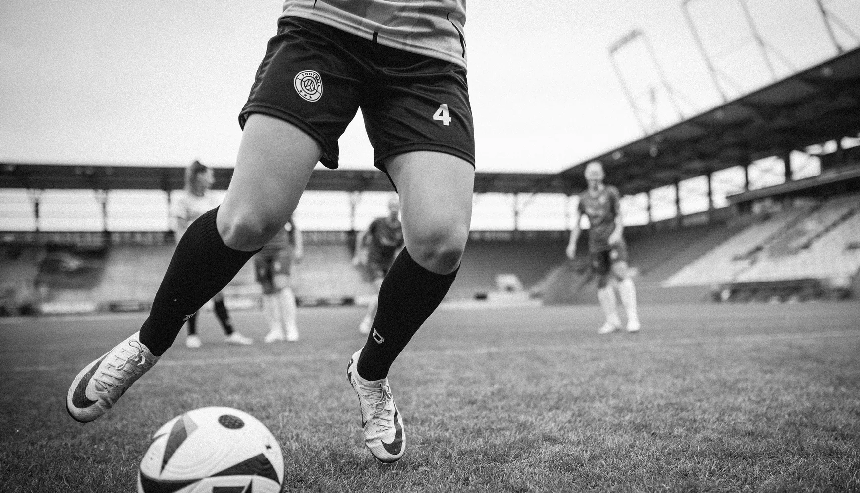A female player in short soccer shorts and long socks is kicking a ball on a soccer field.