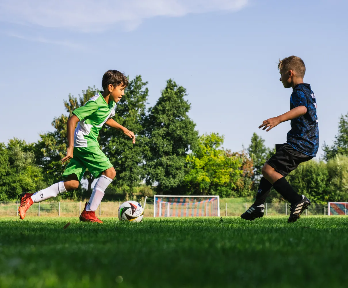 A young soccer player in a green jersey runs with the ball on a grassy field.