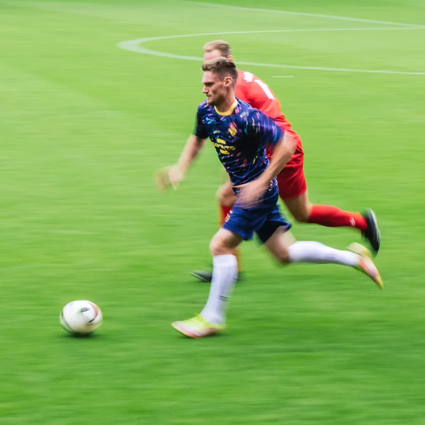 A football player in a colorful jersey sprints across the field while another player chases him.