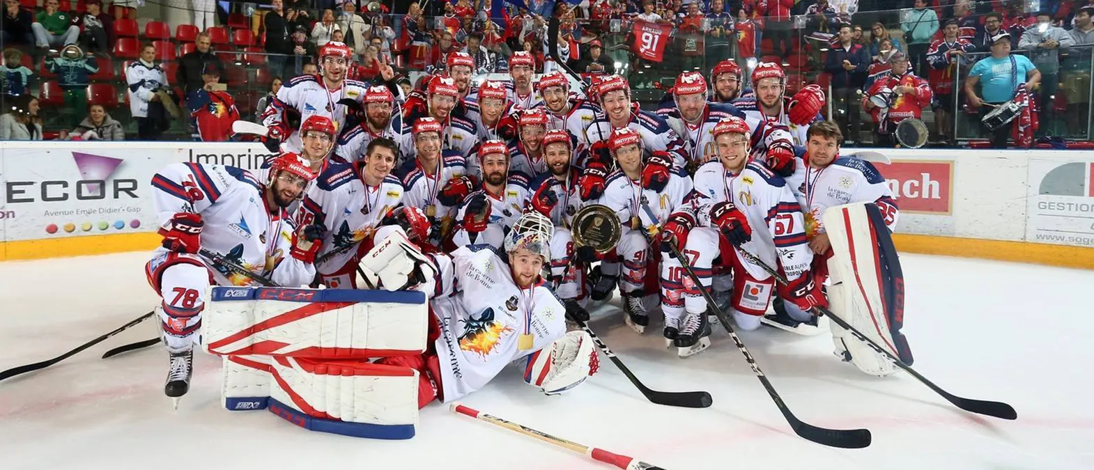 A hockey team in white jerseys with red and blue accents poses on the ice, surrounded by fans, as players wear medals and hold a trophy.