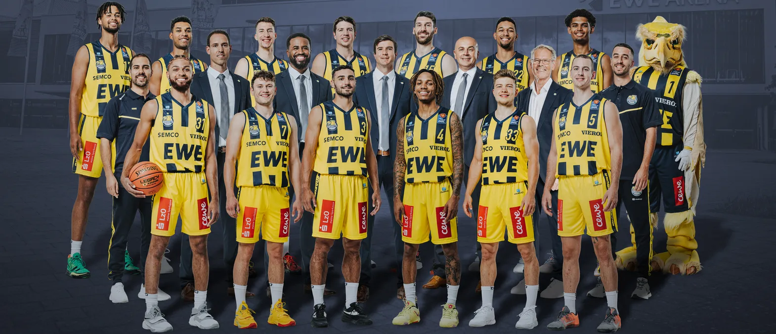 A group photo of a basketball team in yellow and black jerseys with the sponsors 'EWE' and 'SEMC', a mascot, and a basketball, in front of a building.