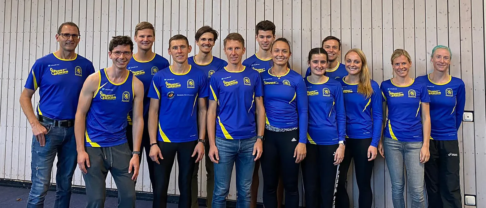 A group of men and women in blue and yellow sportswear with the 'Sport Jakob Passau' logo pose in front of a light wooden wall.