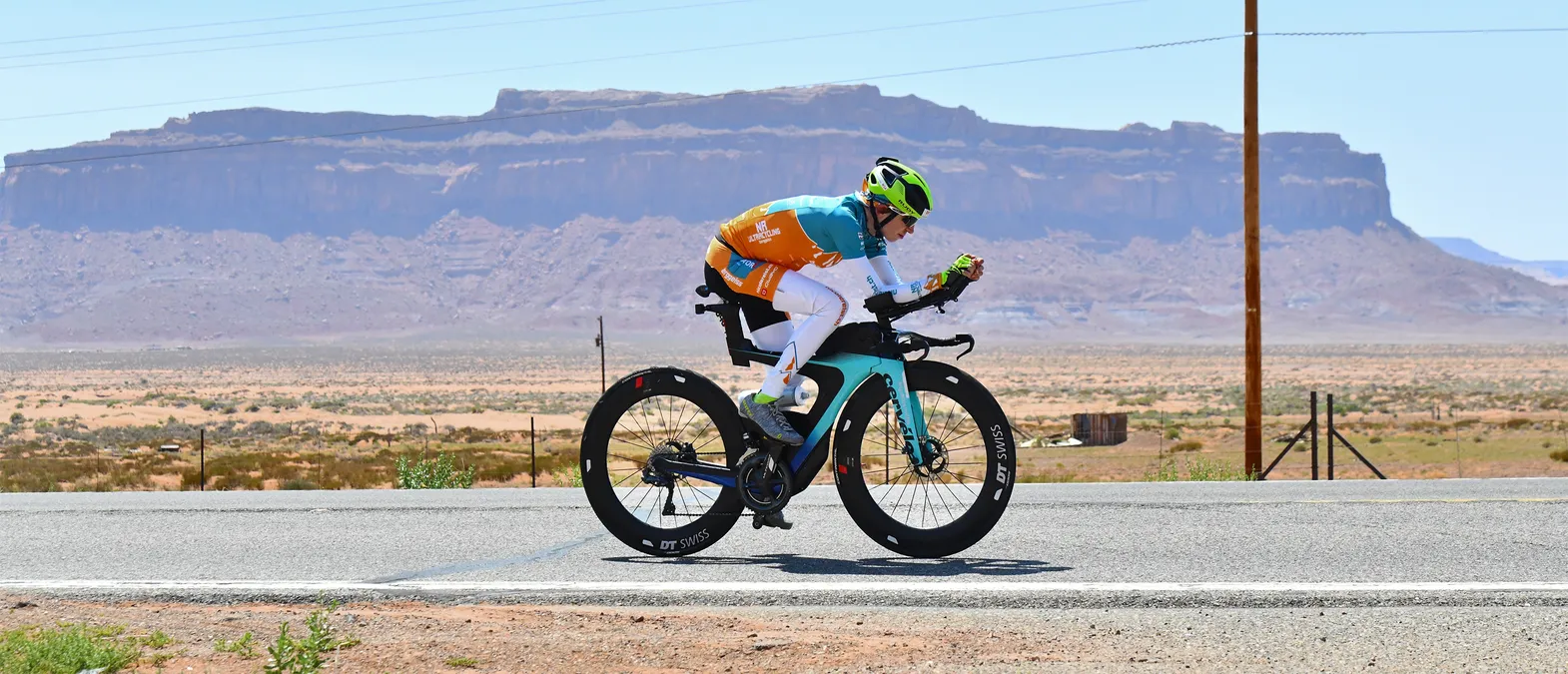 A cyclist in an orange and teal cycling kit riding a time trial bike on a road through a desert landscape with rock formations in the background.