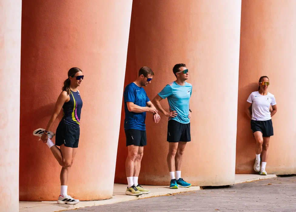 A group of four athletes in running attire stretching against a wall with large columns.