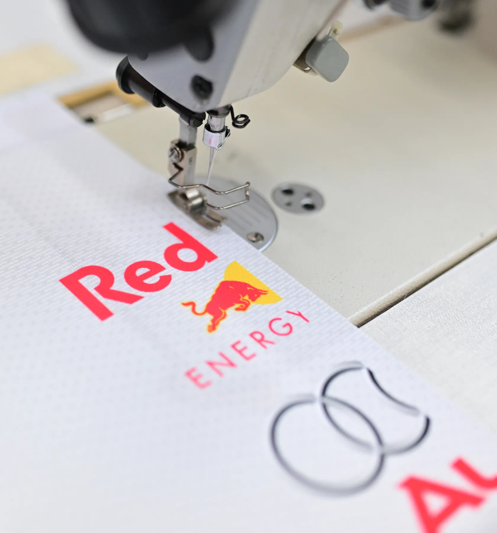 A sewing machine is stitching a white sports garment with a red Red Bull logo.