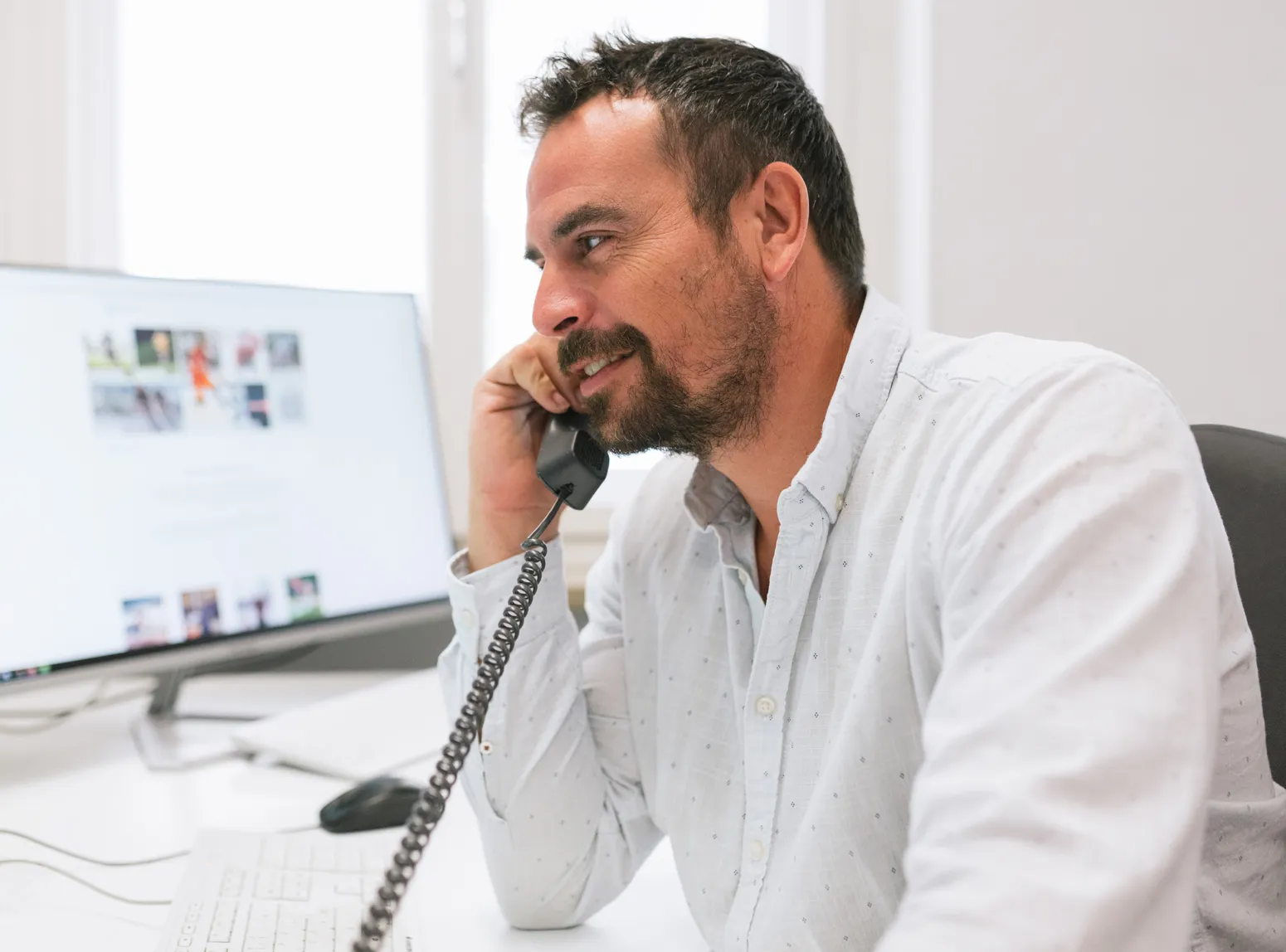 A man with a beard sits at a desk in front of a computer, smiling while talking on an old-fashioned telephone receiver.