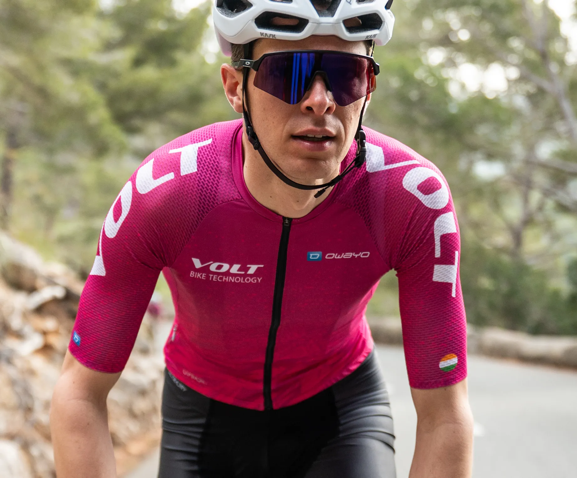 A cyclist wearing a pink short-sleeved cycling jersey with the 'VOLT BIKE TECHNOLOGY' logo and the 'owayo' logo on the chest, riding on a road in a wooded area.