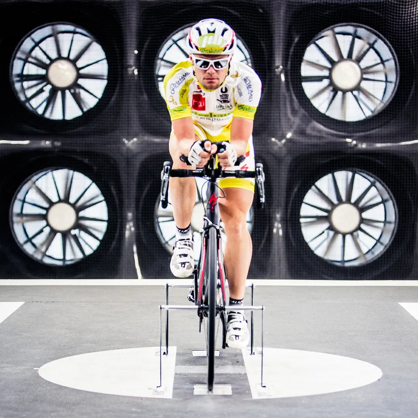 A cyclist training on a stationary bike in a testing lab to optimize aerodynamics, with large fans operating in the background.