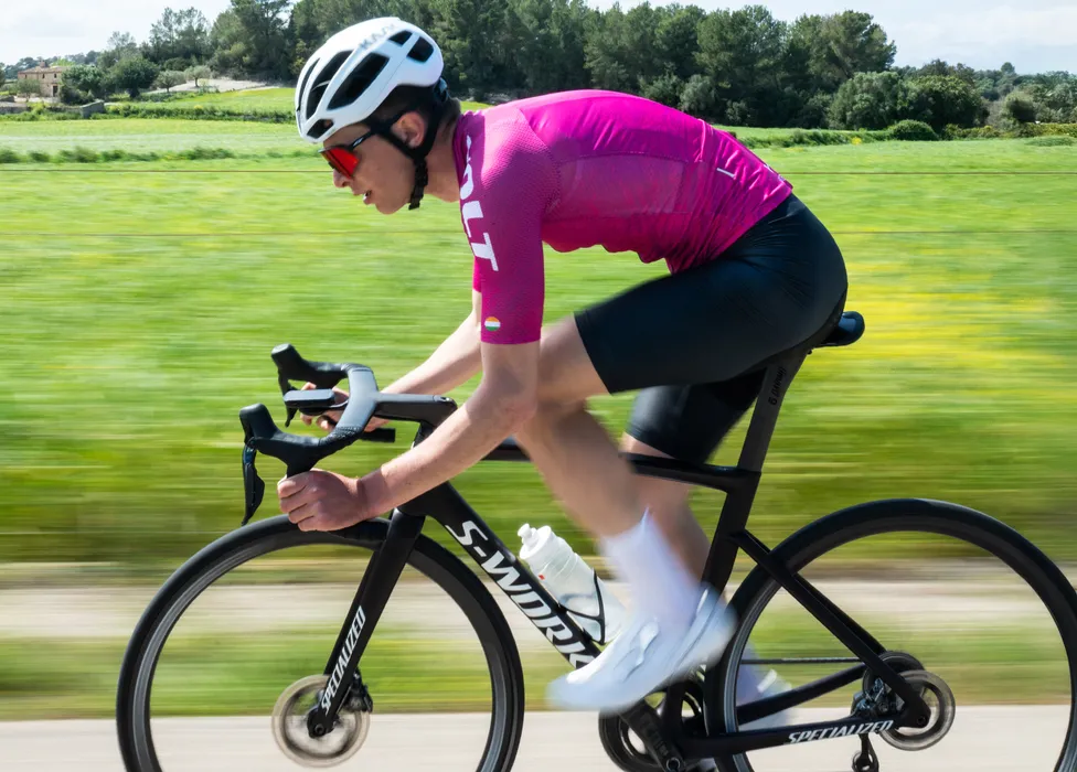 A cyclist in a pink jersey and black shorts rides on a road through a green landscape, with motion blur emphasizing speed.