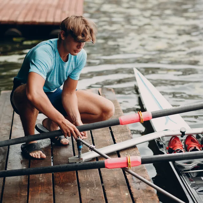 A young man in a blue sports shirt is sitting on a dock assembling oars for a boat, with water and a boat visible in the background.