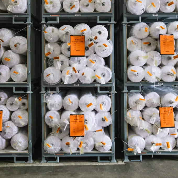 A warehouse with shelves filled with large rolls of white fabric wrapped in plastic and tagged with orange labels.